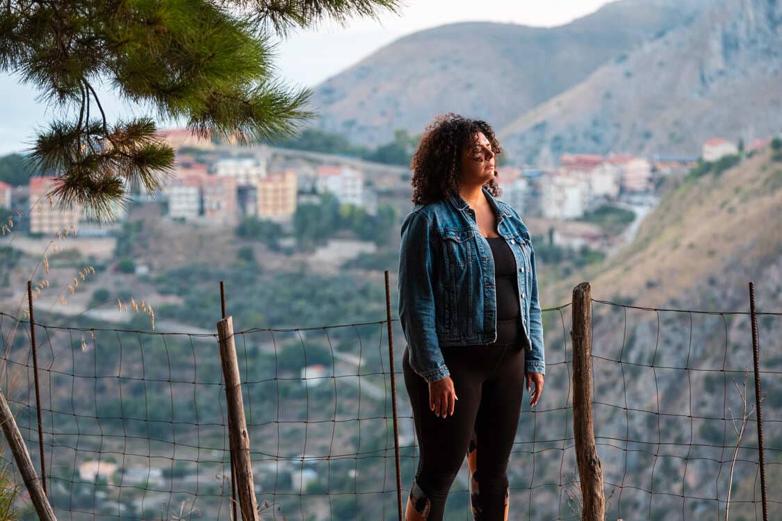 A woman in a denim jacket and black pants stands by a wire fence, with mountains and a village in the background on a cloudy day. Pine tree branches frame the top left corner of the scene.